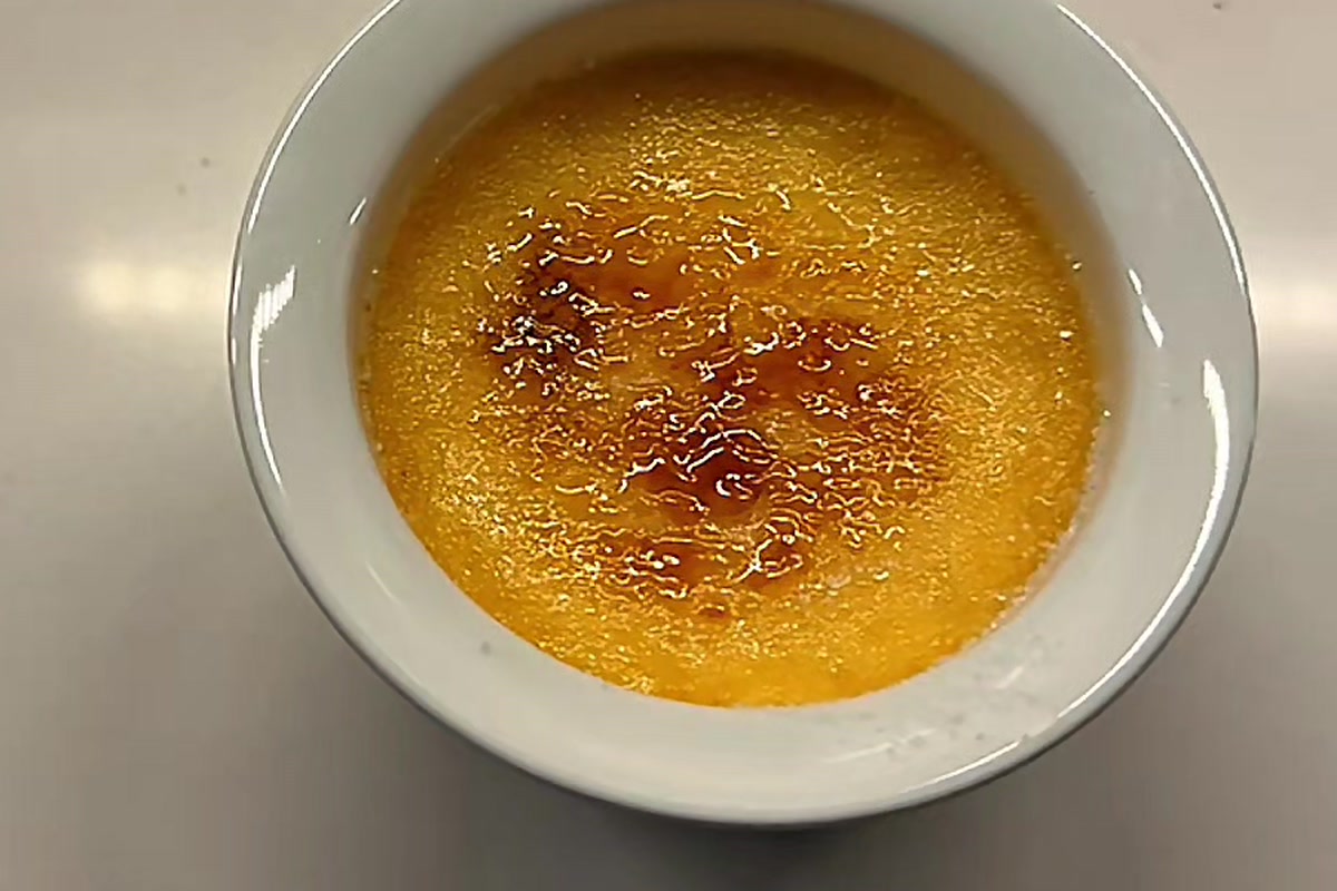 A white ramekin of crème brûlée seen from above, its sugar crust torched to a crackled amber with darker caramel patches across the middle